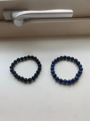 Två pärlarmband i svart och blått - Säljer två stilrena armband med pärlor, ett i mattsvart och ett i blått med marmorerad effekt. Båda är elastiska och passar perfekt för att ge din look en cool touch. Materialet är troligen sten eller glas. Perfekt för dig som gillar minimalistiska accessoarer.