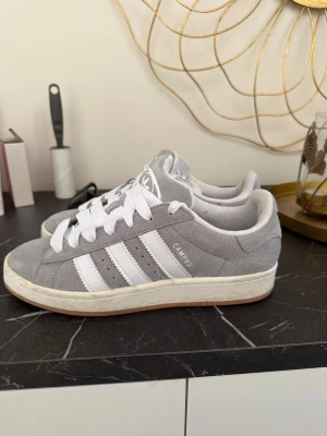 Adidas Campus grå sneakers - Grå Adidas Campus sneakers i mocka med klassiska vita ränder på sidorna och vit logga på hälen. Skorna har vit sula och vita skosnören, samt låg profil som ger en clean och tidlös look. Perfekta för dig som gillar streetstyle och enkel design.