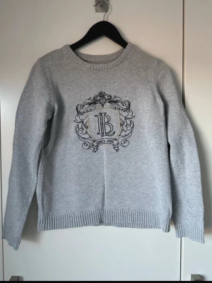 Grå stickad tröja - Snygg grå stickad tröja med rund hals och ett stort broderat emblem med bokstäverna 'LB' och texten 'Since 1986' på bröstet. Tröjan har ribbade muddar vid ärmslut och nederkant, perfekt för en stilren och avslappnad look.