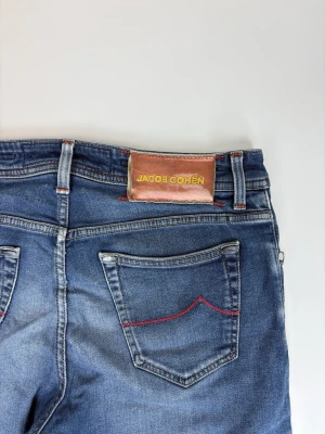 Blå jeans från Jacob Cohën - Snygga blå jeans från Jacob Cohën med klassisk femficksdesign, röda sömmar på bakfickorna och läderpatch med logga. Jeansen har en smal passform och lätt slitning på låret för en trendig look. Perfekta för dig som gillar stilrena detaljer.