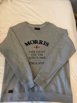 Grå sweatshirt från Morris Gents - Snygg grå sweatshirt från Morris Gents med broderad text och brittisk flagga på bröstet. Tröjan har rund hals, långa ärmar och ribbade muddar. Perfekt för en stilren och avslappnad look. Tillverkad i mjukt bomullsmaterial.