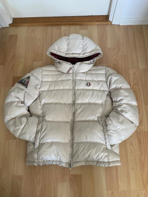 Beige pufferjacka från Dolomite - Snygg beige pufferjacka från Dolomite med broderad logga på bröstet och stor Dolomite-text på ryggen. Jackan har huva, dragkedja framtill och två sidofickor samt två innerfickor. Perfekt för kalla vinterdagar och riktigt skön att bära. Materialet är glansigt och quiltat. Den är lite sketen men väldigt snygg förtfarande.  Det står XL men den är S/M