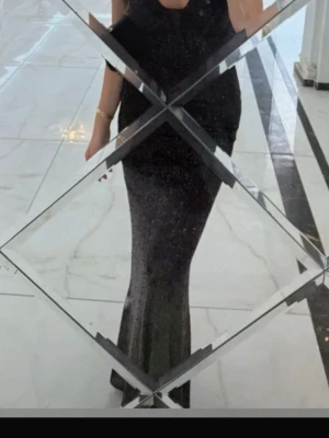 Svart glittrig långklänning - Svart långklänning med smala axelband och djup v-ringning. Klänningen är täckt av glittriga paljetter och har en figurnära passform som framhäver siluetten. Perfekt för dig som vill sticka ut med en elegant och glamorös look.