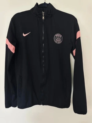 Svart/rosa PSG dress från Nike - Snyggt svart träningsset från Nike och Paris Saint-Germain med rosa detaljer. Jackan har dragkedja, ståkrage och PSG-logga på bröstet. Byxorna har matchande rosa rand och PSG-märke. Perfekt för fotbollsträning eller chill.