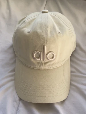Offwhite keps från Alo - Snygg beige keps från Alo med broderad logga framtill. Klassisk böjd skärm och justerbar rem baktill. Tillverkad i mjuk bomull för en bekväm passform. Perfekt accessoar för en avslappnad och trendig look.