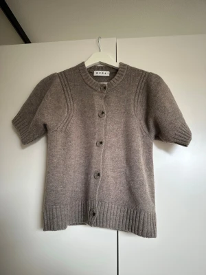 Beige stickad kofta från Wera - Kortärmad stickad kofta i beige från Wera. Koftan har rund halsringning, ribbstickade muddar och knäppning framtill med fem knappar. Mjuk och skön ullblandning, perfekt för lager-på-lager-stil.