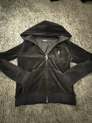 Svart velour hoodie Juicy Couture - Snygg svart hoodie i mjuk velour från Juicy Couture. Dragkedja framtill, två fickor och klassisk huva. Liten JC-logga broderad på bröstet. Perfekt för en chill och trendig look.