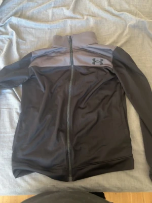 Svart träningsjacka från Under Armour - Snygg svart träningskofta från Under Armour med grå panel upptill och dragkedja hela vägen. Koftan har hög krage och är tillverkad i ett stretchigt, funktionsmaterial som andas. Perfekt för träning eller uppvärmning.