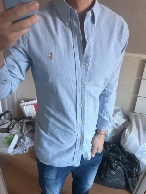 Ljusblå skjorta från Ralph Lauren - Klassisk ljusblå skjorta från Ralph Lauren med button-down krage och broderad logga på bröstet. Skjortan har långa ärmar och knäppning framtill. Perfekt för en stilren och avslappnad look.