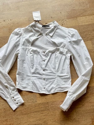 Vit statementblus från Mohito - Trendig vit blus från Mohito med unik cut-out detalj vid bröstet och markerad krage. Blusen har långa ärmar med knappar vid ärmslut och figurnära passform. Perfekt för dig som vill sticka ut med en modern twist på klassisk skjortstil.