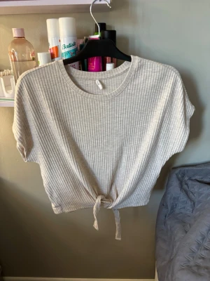 Beige ribbad topp med knyt framtill - Superfin beige ribbad topp från Only med korta ärmar och knyt framtill. Toppen har en avslappnad passform och är tillverkad i mjukt material som känns skönt mot huden. Perfekt att styla med jeans eller kjol för en trendig look.