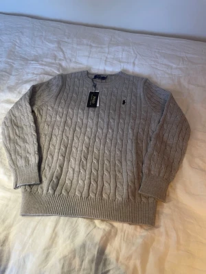 Ljusgrå kabelstickad tröja Polo Ralph Lauren - Kabelstickad ljusgrå tröja från Polo Ralph Lauren med rund halsringning och klassisk broderad logga på bröstet. Tröjan har ribbade muddar vid ärmslut och nederkant, perfekt för en stilren och avslappnad look. Storlek S. Taggen man fick när ja köpte den finns även kvar som syns på bilderna.