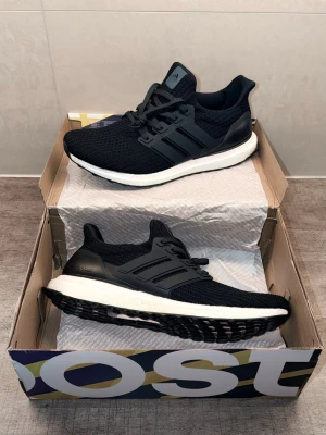 Adidas Ultraboost svarta sneakers - Svarta Adidas Ultraboost sneakers med stickad ovandel, klassiska tre ränder på sidan och vit Boost-mellansula. Yttersulan har ett unikt mönster för extra grepp. Skorna har en sportig och modern look med låg profil och snörning.