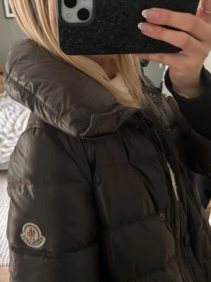 Brun pufferjacka från Moncler - Säljer en brun pufferjacka från Moncler med hög krage och quiltad design. Jackan har dragkedja och tryckknappar framtill samt Moncler-logga på ärmen. Perfekt för kalla dagar och riktigt snygg till alla outfits. Köpt för 3000 kr på vestiare collective 