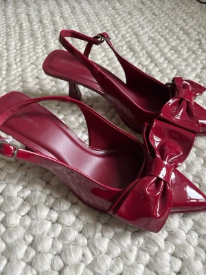 Vinröda lackpumps med rosett - Snygga vinröda pumps i lack med spetsig tå och stor dekorativ rosett framtill. Skorna har öppen häl med justerbar rem och en smal klack. Perfekta för dig som vill sticka ut med en trendig och elegant look. Använda en gång 