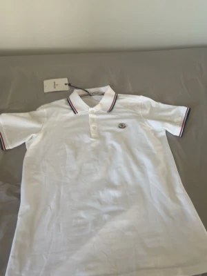 Vit pikétröja från Moncler - Vit pikétröja från Moncler. Det är storlek L men passar och sitter som M. Den är för liten för folk med L. Den har en klassisk krage och knäppning framtill. Tröjan har röda och blå detaljer på krage och ärmslut samt Moncler-logga på bröstet. Perfekt för en stilren och sportig look. 