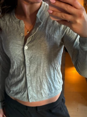 Grå ribbad kofta med knappar - Snygg grå kofta med långa ärmar och ribbade muddar. Modellen är croppad och har knäppning framtill med vita knappar. Perfekt att slänga över en topp för en chill look.