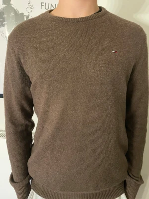 Brun stickad tröja från Tommy Hilfiger - Snygg brun stickad tröja från Tommy Hilfiger med rund halsringning och diskret logga på bröstet. Tröjan har långa ärmar och ribbade muddar vid ärmslut och nederkant. Perfekt för lager-på-lager och enkel att matcha med jeans eller chinos.