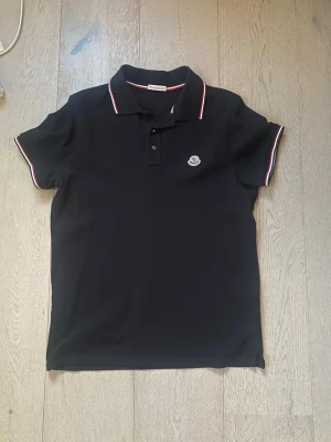 Svart pikétröja från Moncler - Klassisk svart pikétröja från Moncler med korta ärmar och vit-röda detaljer på krage och ärmslut. Tröjan har knappslå framtill och Moncler-logga på bröstet. Perfekt för en stilren och sportig look.