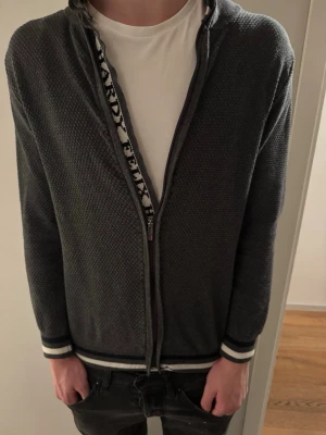 Grå ziptröja från Felix Hardy - Snygg grå ziptröja från Felix Hardy med diskret strukturmönster. Tröjan har hel dragkedja, ribbade muddar och nederkant med vita och svarta ränder. Perfekt att slänga på sig över en t-shirt för en avslappnad look.