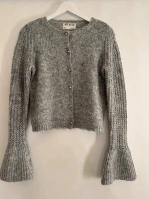Grå stickad kofta från AWARE - Mysig grå stickad kofta från AWARE med rund halsringning och pärlknappar framtill. Koftan har långa ärmar med utsvängda muddar och en mjuk, fluffig känsla. Perfekt för lager på lager och en trendig look. 