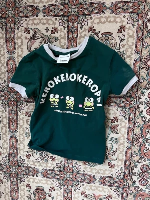 Grön Keroppi t-shirt med tryck - Säljer en grön Keroppi t-shirt från Bershka med vitt tryck och söta grodor framtill. T-shirten har vita detaljer vid ärmslut och hals, är kortärmad och croppad modell. Perfekt för dig som gillar gulliga motiv och vill sticka ut lite extra.