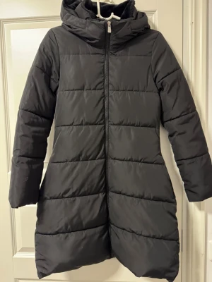 Svart lång dunjacka med huva - Snygg svart lång dunjacka med huva och dragkedja framtill. Jackan har en stilren design med horisontella sömmar och är perfekt för kalla vinterdagar. 