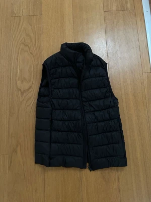 Svart dunväst  - Snygg svart dunväst med quiltad design och ståkrage. Västen har dragkedja framtill och är lätt vadderad för extra värme. Perfekt att slänga över en hoodie eller tröja för en cool lager-på-lager-look.