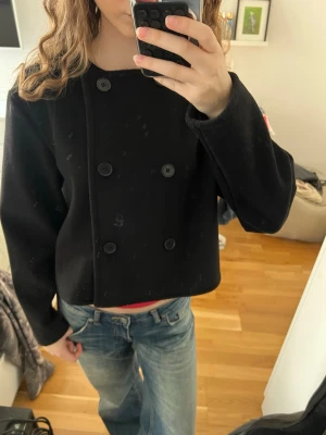 Svart skepparkavaj med dubbelknäppning - Stilren svart skepparkavaj med kort, boxig passform och klassisk dubbelknäppning framtill. Jackan har rundad krage och är tillverkad i ett tjockare tyg som ger en snygg siluett. Perfekt för dig som gillar minimalistisk och tidlös stil.