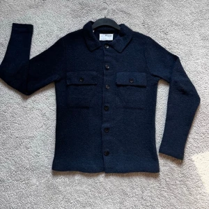 Mörkblå overshirt från Selected Homme - Stilren mörkblå overshirt från Selected Homme med två stora bröstfickor med knappar och klassisk krage. Tillverkad i ett mjukt, strukturerat material som ger en avslappnad men snygg look. Perfekt att bära öppen eller stängd över en t-shirt.