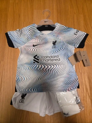 FC Liverpool Nike fotbollsset barn unisex  strl. XS96-104 cm - Säljer ett Liverpool FC bortaställ från Nike med tröja, shorts och strumpor. Tröjan har ett unikt mönster i vitt, blått och svart med klubbmärke och sponsortryck. Korta ärmar och rund hals. Perfekt för fotbollsträning eller match.