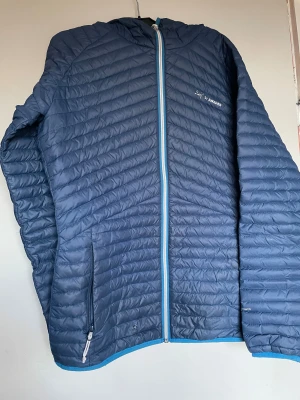 Blå dunjacka från 2117 of Sweden - Snygg blå dunjacka med huva och dragkedja i kontrasterande ljusblå färg. Jackan har två fickor med dragkedja och quiltad design. Riktigt snygg blåfärg. I bra skick förutom lilla hålet som är lagat med genomskinligt nagellack😊
