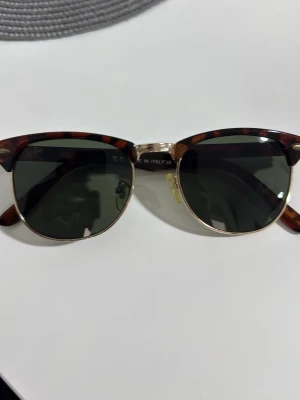Ray-Ban Clubmaster solglasögon brun - Säljer ett par klassiska Ray-Ban Clubmaster solglasögon med brunmelerad båge och guldfärgade detaljer. Glasen är mörkgröna och bågen har en retroinspirerad form. Tillverkade i Italien och perfekta för dig som vill ha en tidlös look.