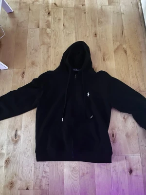 Svart hoodie storlek m/s - Svart hoodie med dragkedja. Klassisk design med huva och dragsnören, samt den ikoniska vita broderade loggan på bröstet. Perfekt för en avslappnad och stilren look.