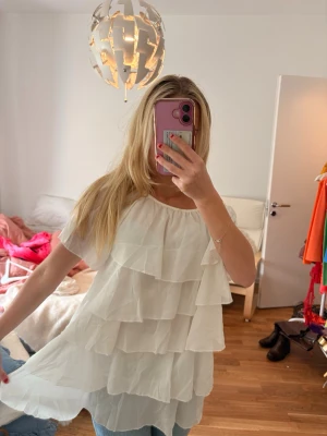 Vit volangblus med kort ärm - Säljer en söt vit blus med flera lager volanger och korta ärmar. Blusen har en lös passform och är tillverkad i ett lätt, luftigt material som ger ett flowy intryck. Perfekt att styla med jeans eller kjol för en trendig look. (Liten fläck i fram, går bort i tvätt med lite vanilj)
