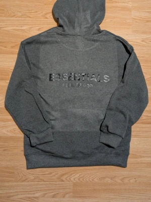 Grå hoodie Essentials Fear of God - Snygg grå hoodie från Essentials Fear of God med broderad logga på bröstet. Klassisk passform med huva och ribbade muddar. Perfekt för en avslappnad och trendig stil. Mjuk bomullsblandning som känns skön mot huden.