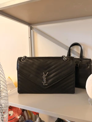 Svart YSL axelväska i läder - Snygg svart axelväska från Saint Laurent i quiltat läder med silverfärgade kedjor och ikoniska YSL-loggan framtill. Väskan har ett klassiskt kuvertformat lock och stilren design som passar till det mesta. Perfekt för dig som vill ha en lyxig och tidlös accessoar.