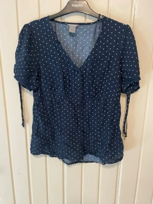 Blå prickig blus från Lindex - Mörkblå blus från Lindex med vita prickar, v-ringning och knappar framtill. Blusen har korta ärmar med rynkade detaljer och knytband. Tunt och luftigt material, perfekt för varmare dagar.