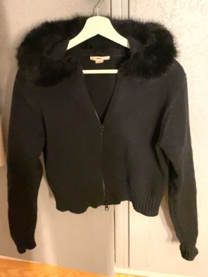 Svart cardiganjacka med fuskpäls - Svart stickad cardiganjacka från Gina Tricot med dragkedja och huva dekorerad med fuskpäls. Det går även att knäppa av pälsen om man inte vill ha den. Jackan är  perfekt för en trendig och mysig look.
