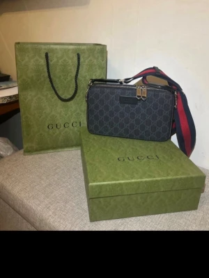 Gucci axelväska med monogram - Säljer en exklusiv Gucci crossbody bag i svart/mörkgrå GG Supreme canvas med silverdetaljer och svart/röd justerbar axelrem. Väskan är endast använd 2 gånger och är i nästintill nyskick.  Köpt direkt i Gucci butik för 14 500 kr. Originalkvitto, box, Gucci-påse och samtliga tillbehör medföljer.  En stilren och tidlös designerbag med flera praktiska fack – perfekt både till vardag och finare tillfällen.  Endast seriösa köpare. Pris diskuteras!