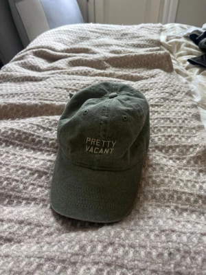Olivgrön keps med text Pretty Vacant - Snygg olivgrön keps med broderad vit text 'PRETTY VACANT' framtill. Klassisk böjd skärm och justerbart spänne baktill för perfekt passform. Tillverkad i mjukt bomullstyg, perfekt för en avslappnad streetstyle-look.