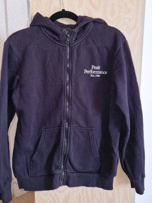 Svart hoodie Peak Performance - Svart hoodie med dragkedja från Peak Performance. Klassisk modell med huva, två fickor framtill och logga broderad på bröstet. 