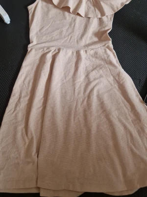 Beige oneshoulder klänning med volang - Snygg beige/ rosa  klänning med volangdetalj över axeln. Klänningen har en mjuk, lätt struktur och är perfekt för varma dagar. Den har en avslappnad passform och är tillverkad i ett bekvämt material. Har glitter 