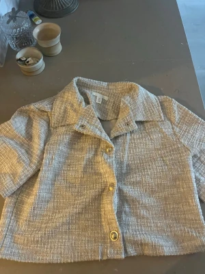 Beige kortärmad blus med struktur - Stilren beige blus från H&M med korta ärmar och markerad krage. Blusen har en strukturerad väv och knäpps framtill med tre knappar i guldton. Perfekt för en trendig och avslappnad look.
