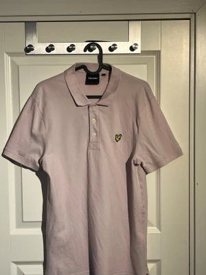 Ljuslila pikétröja från Lyle & Scott - Snygg ljuslila pikétröja från Lyle & Scott med klassisk krage och tre knappar framtill. Tröjan har korta ärmar och den ikoniska gula loggan broderad på bröstet. Perfekt för en stilren och avslappnad look.