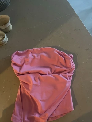 Rosa axelbandslös kort topp - Säljer en söt rosa axelbandslös kort klänning med draperad detalj upptill. Perfekt för dig som gillar en enkel men snygg look. Klänningen har en mjuk känsla och passar till många olika tillfällen.