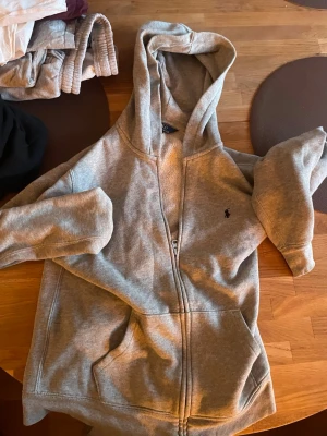 Grå hoodie från Polo Ralph Lauren - Snygg grå zip-hoodie från Polo Ralph Lauren med klassisk huva och dragkedja framtill. Ikoniska lilla loggan broderad på bröstet. Mjuk insida och två fickor framtill. Perfekt för en chill och avslappnad stil.