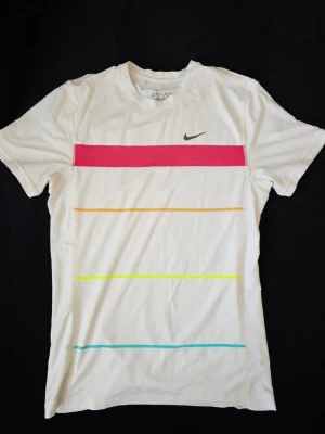 Vit Nike tenniströja, Nadal edition - Vit t-shirt från Nike med korta ärmar och färgglada horisontella ränder i rosa, orange, gul och blått. Liten Nike-logga på bröstet. Tillverkad i ett lätt och sportigt material, perfekt för en aktiv livsstil. 