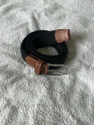 Svart flätat bälte med bruna detaljer - Stilrent svart flätat bälte med bruna skinn-detaljer och silverfärgat spänne. Perfekt accessoar för att lyfta din outfit med en klassisk touch. Passar till både jeans och kostymbyxor.
