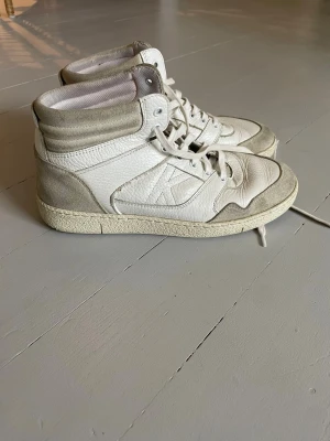 Vita high top sneakers från Ikks - Snygga vita high top sneakers från Ikks (Le Coq Sportif) med detaljer i ljusgrå mocka och vitt läder. Klassisk retrodesign med högt skaft, vita skosnören och diskret logga på sidan. Köpta i London. Säljer pga att mina fötter blivit större.